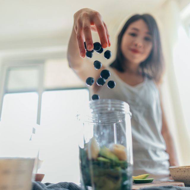 low angle view of a beautiful young asian woman throwing blueberry into the blender to make a healthy smoothie with banana, spinach and vegan milk
