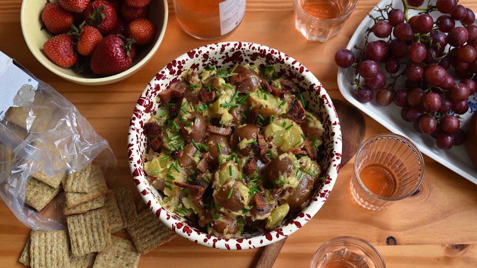 A bowl of potato salad beside glasses of pink wine, crackers, strawberries and grapes