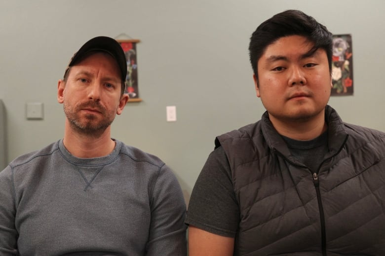Two men pose for a photograph. On the left, a man with a blue long-sleeve shirt who is wearing a hat looks at the camera. On the right, a man wearing a vest and a grey t-shirt looks at the camera.